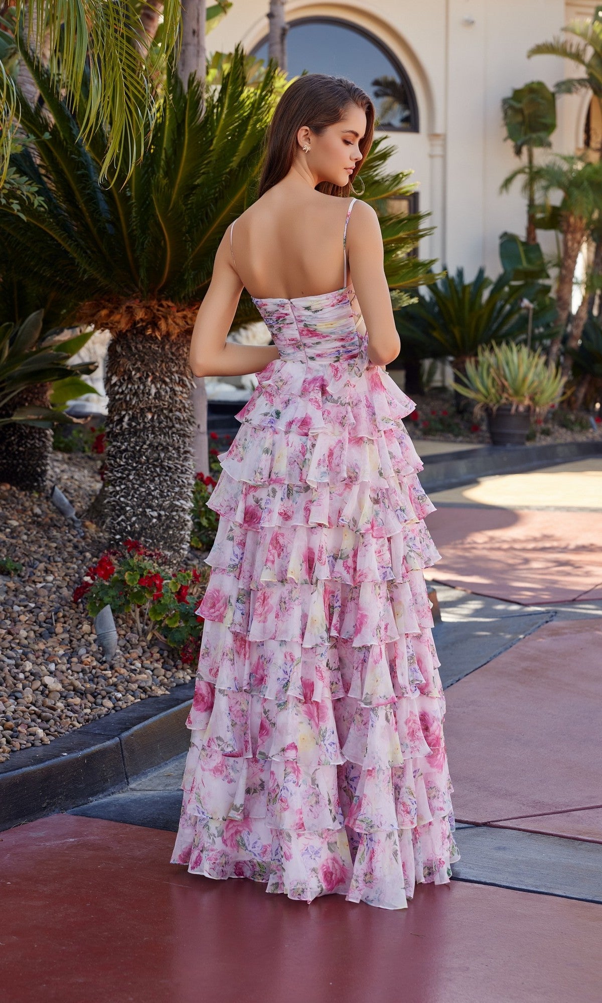 A woman stands outdoors, facing away, in the floor-length Nox Anabel Q1777 prom dress featuring sleeveless floral chiffon, multiple ruffled tiers, and an elegant a-line skirt.